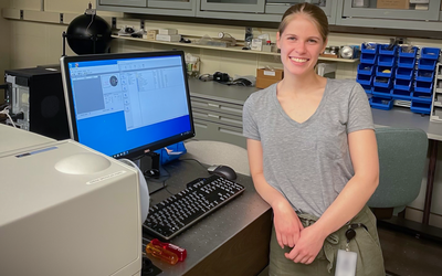 Grace Waters poses smiling in the lab, leaning against a desk with a computer monitor showing data and graphs. 