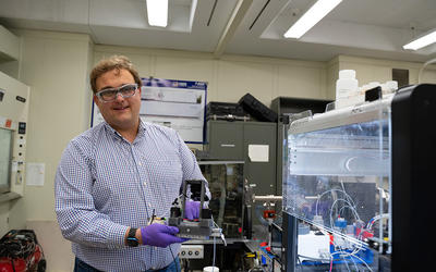 Peter Beaucage wears safety glasses as he stands in the lab, holding a piece of equipment on display. 